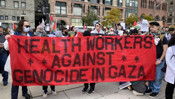 People in a crowd carrying a banner which reads "Health Workers Against Genocide In Gaza".